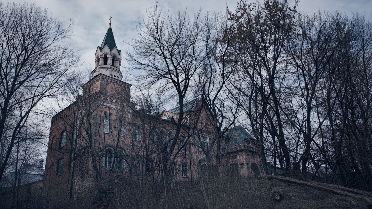 A rusted iron gate in a decaying brick wall under a cold, overcast sky, with a long dirt path leading toward the asylum.